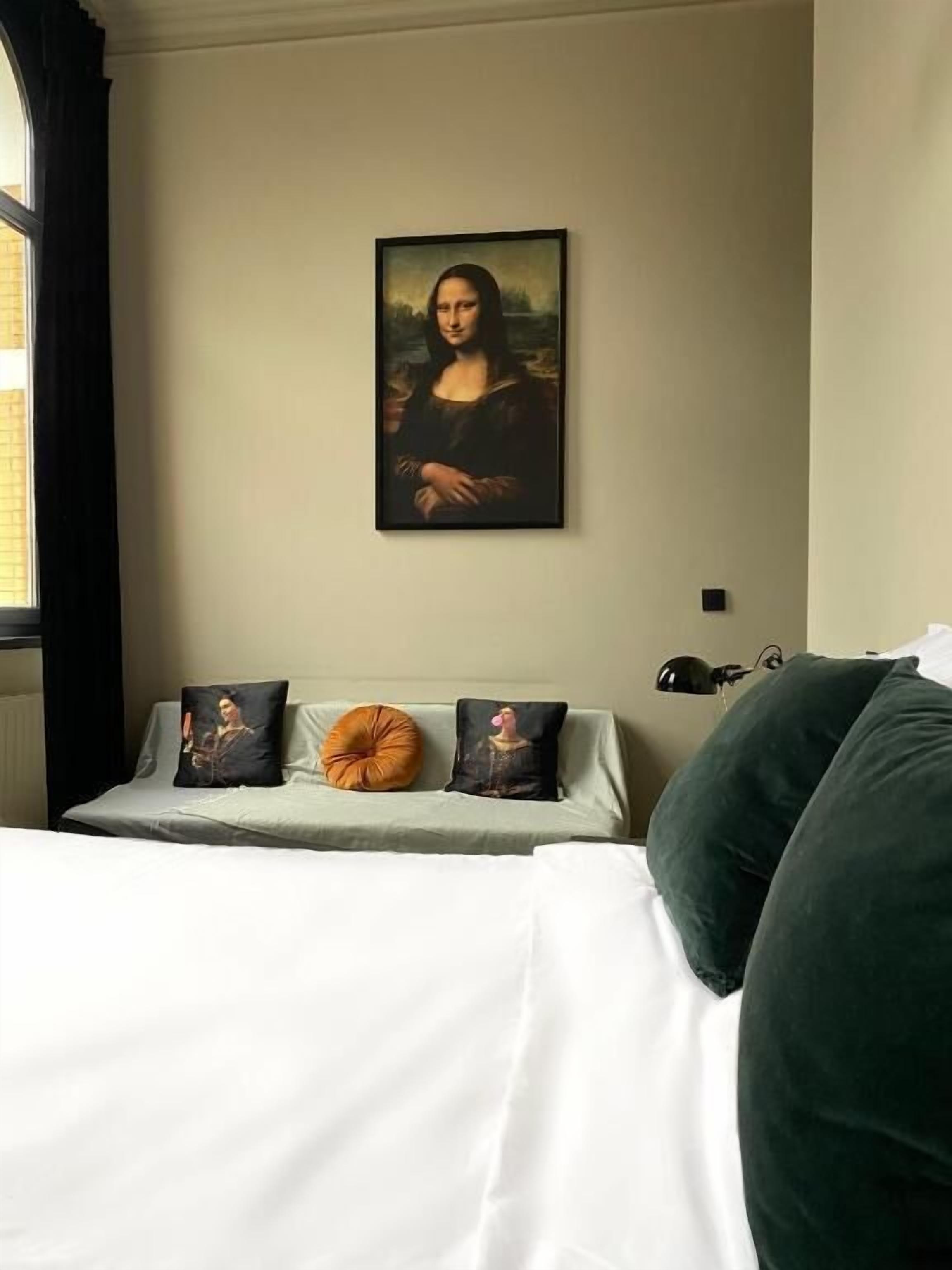 Una habitación con una cama, almohadas, un cuadro colgado en la pared y una ventana con cortinas.
