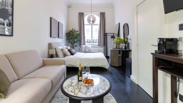 A modern hotel room with a sofa, a bed, a dining table with a bottle and glasses, a small kitchen area, and a large window with curtains.