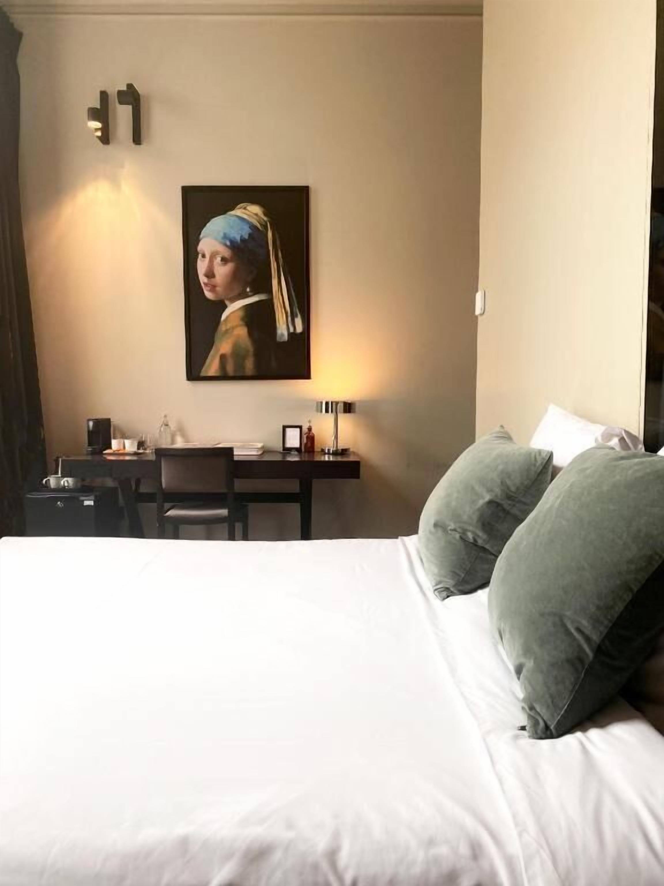 Una habitación de hotel con cama, escritorio, silla, lámpara y un cuadro en la pared.