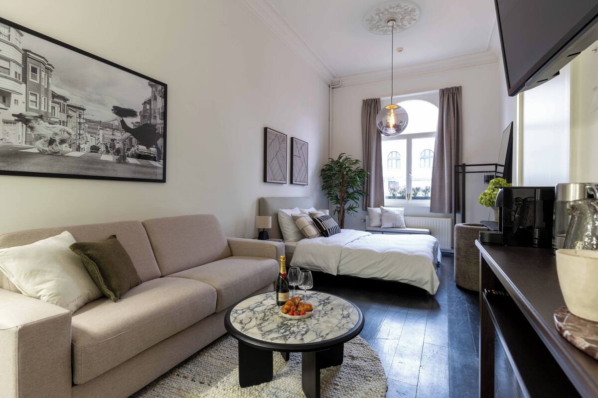 A modern living room with a sofa, two beds, a coffee table with a bottle and glasses, a TV, and a large framed picture on the wall.