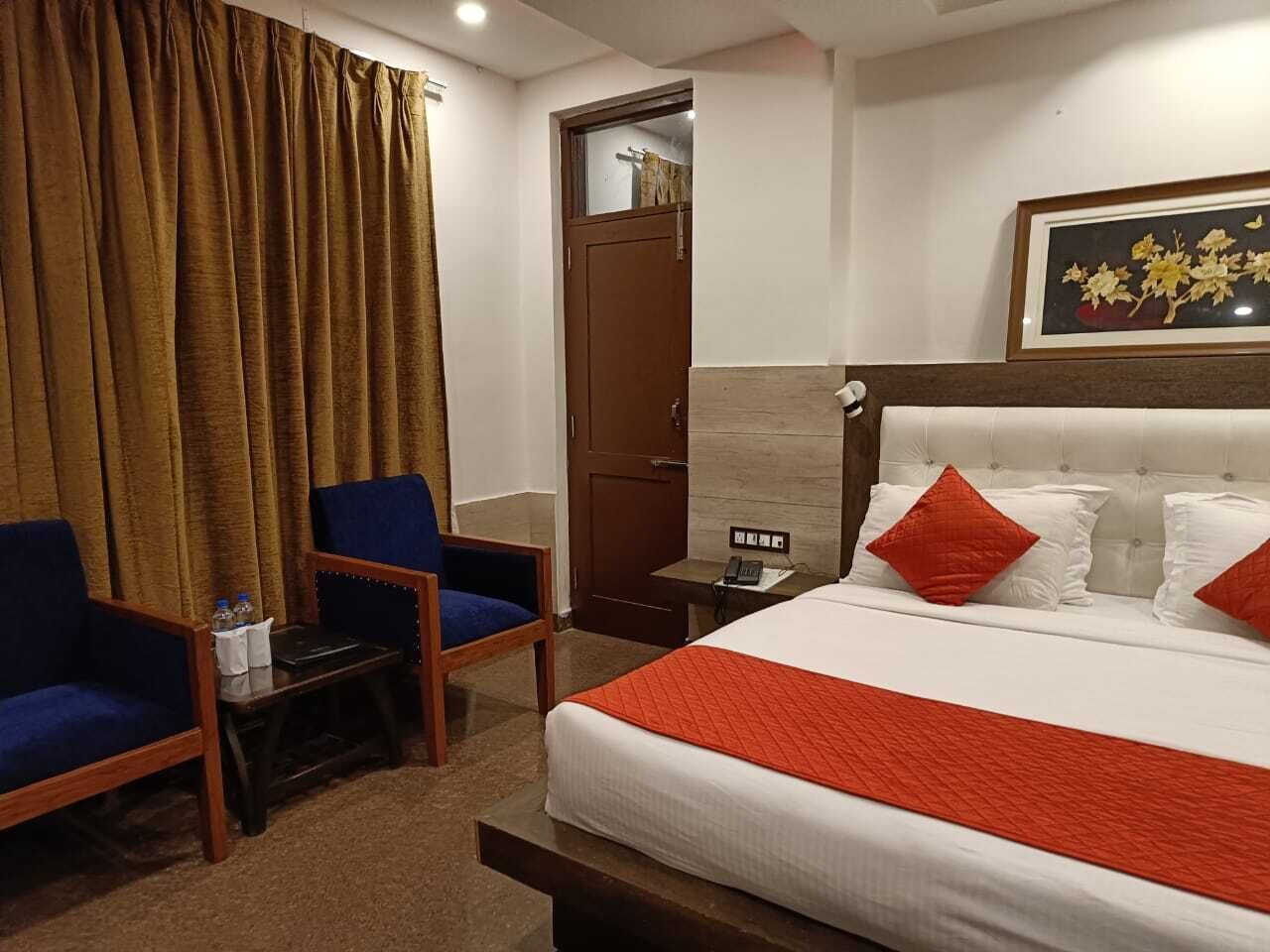 A hotel room with a bed, two blue armchairs, a small table, a nightstand, and a door.