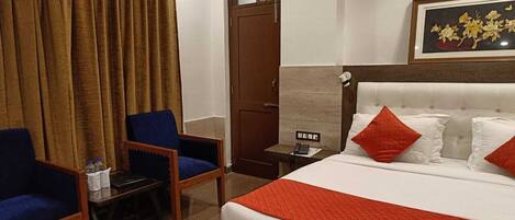 A hotel room with a bed, two blue armchairs, a small table, a nightstand, and a door.