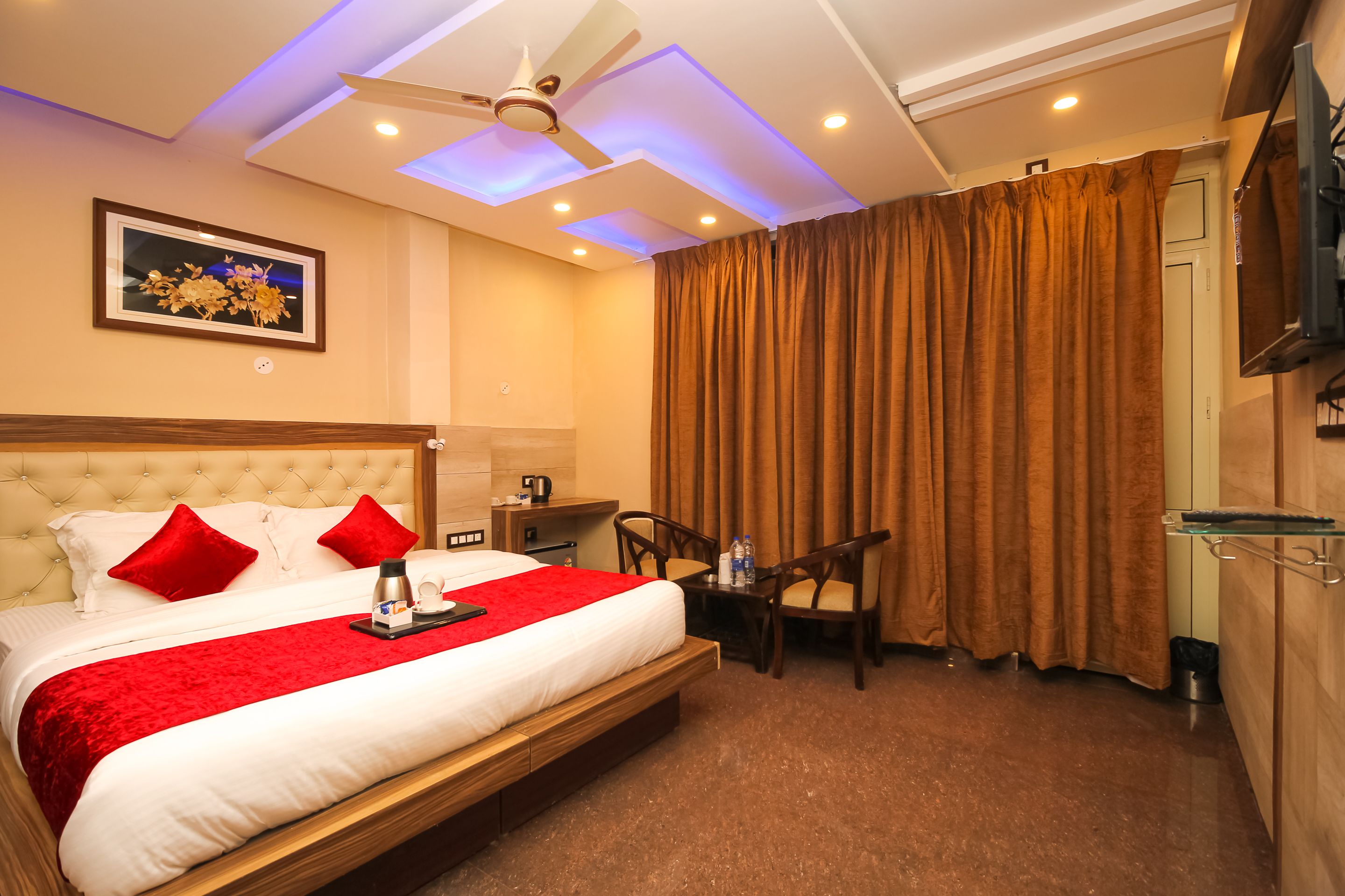A hotel room with a large bed, red pillows, a ceiling fan, a small table with chairs, and a TV on the wall.