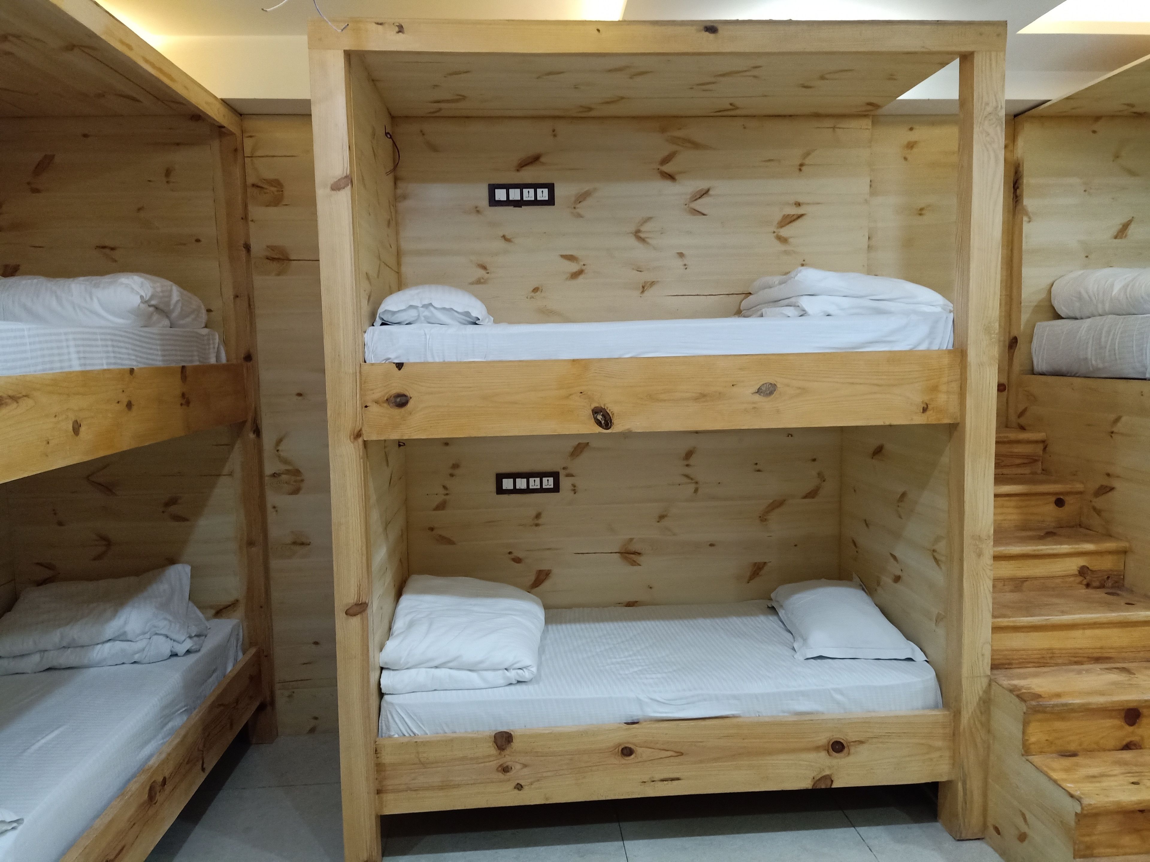 Habitación con literas, dos camas con ropa blanca y un pequeño estante cada una. Hay enchufes eléctricos en la pared sobre las camas.