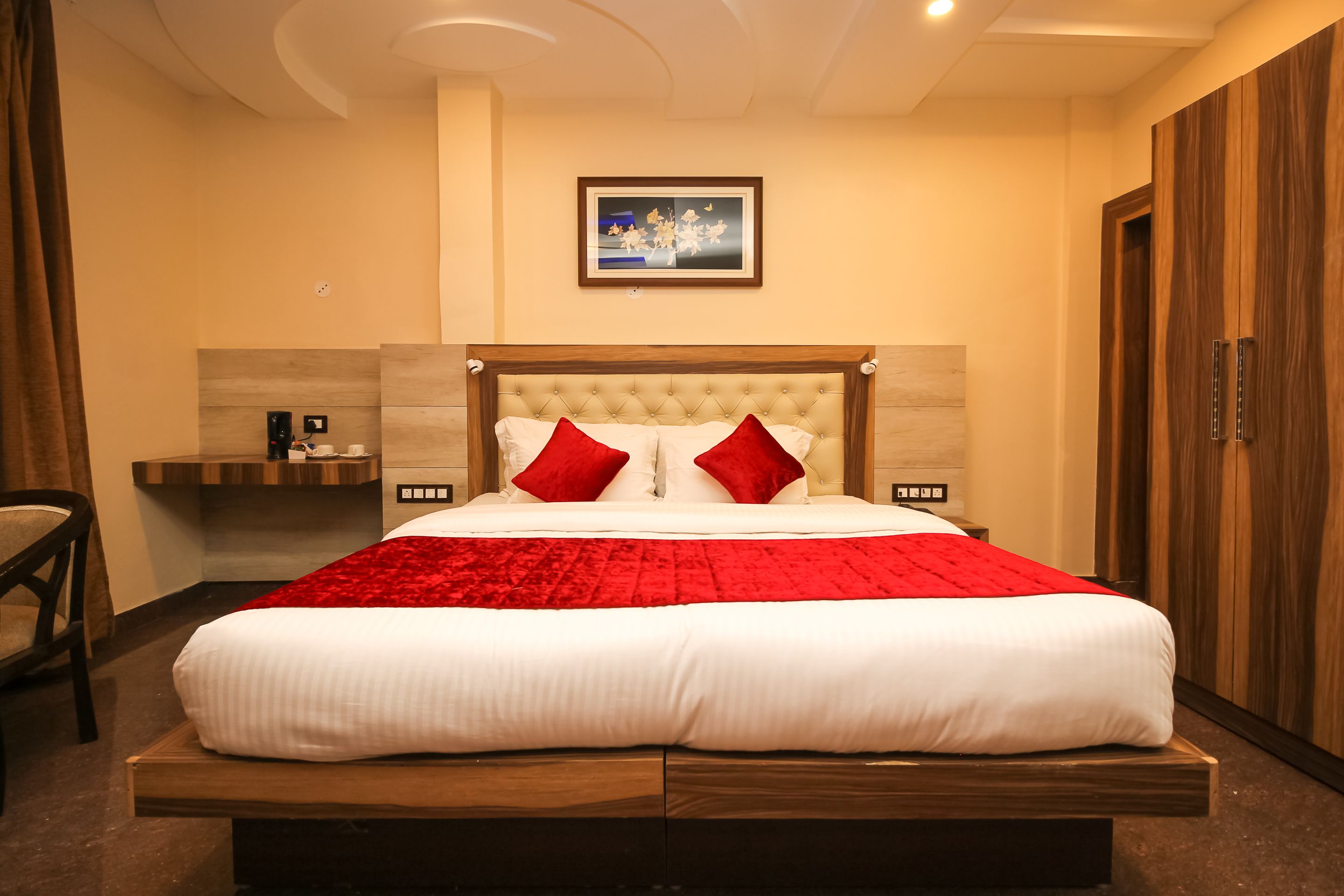Un grand lit avec un cadre en bois, une couvre-lit rouge et blanche et deux oreillers rouges. Une petite table avec une lampe et un cadre photo sont placés sur le mur au-dessus du lit.