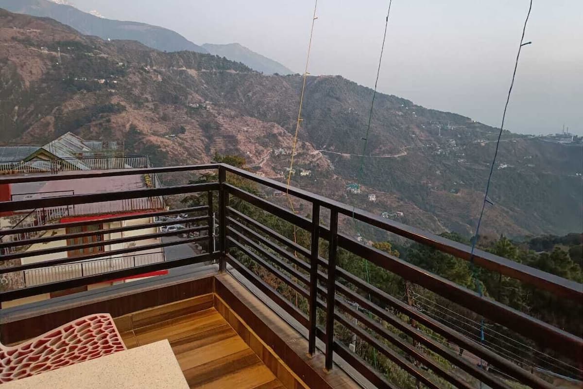 A balcony with a wooden floor and metal railing, overlooking a mountainous landscape with buildings in the distance.