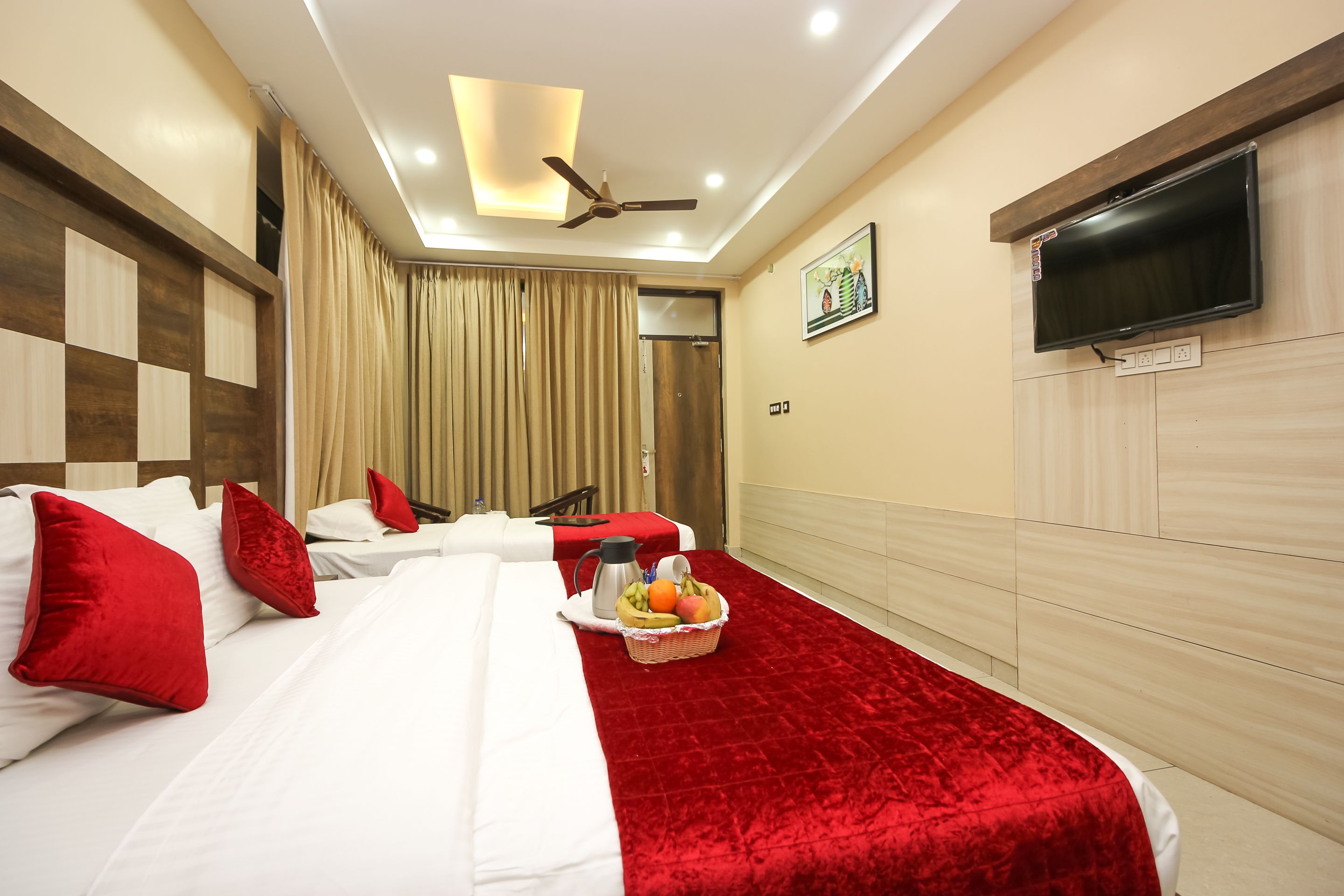 A hotel room with two beds, red and white bedding, a red throw blanket, a fruit basket, a coffee maker, and a flat-screen TV mounted on the wall.