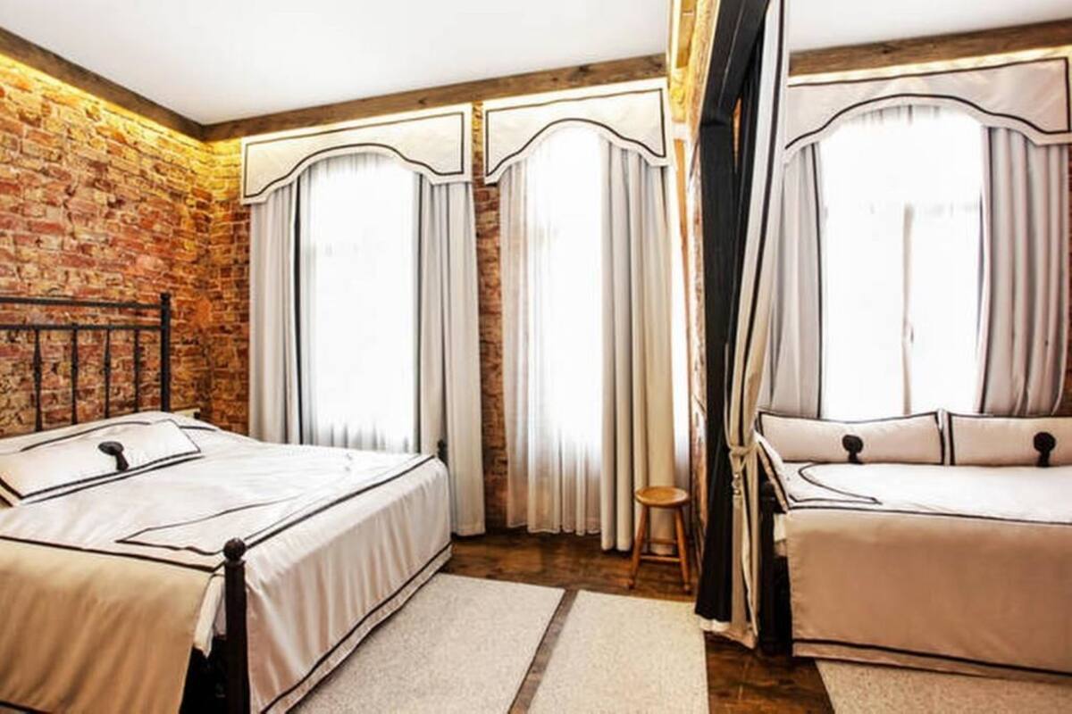 A bedroom with a brick wall, two beds with white bedding, and large windows with curtains.