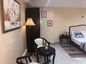 A hotel room with a bed, a desk with a chair, a lamp, and framed pictures on the wall.