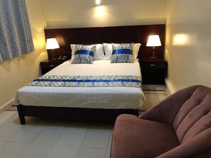 A neatly made bed with patterned bedding, two bedside lamps, a dark headboard, and a brown armchair.