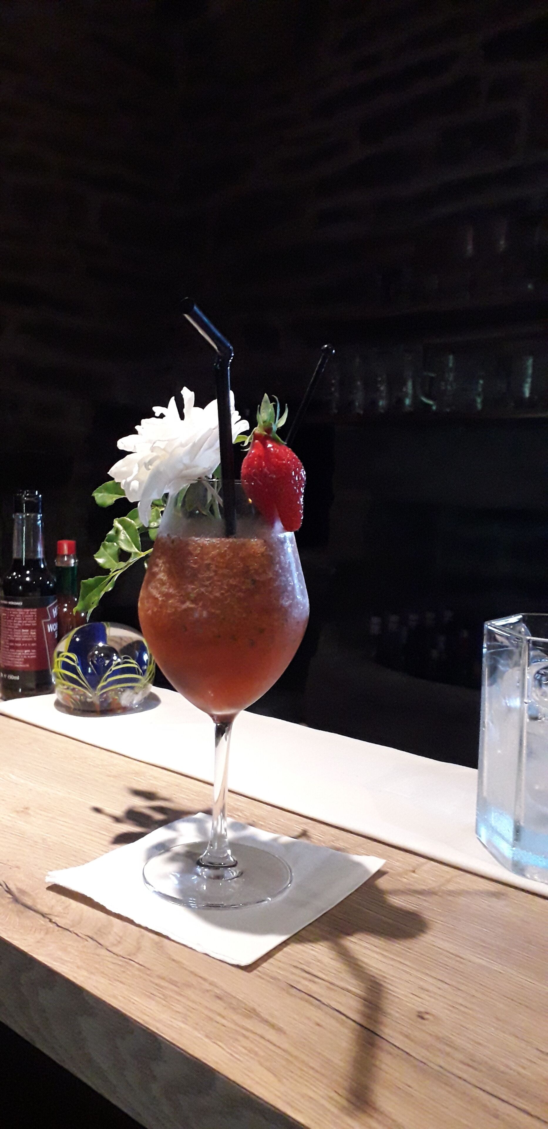 En cocktail med jordbærgarnityr og en blomst, placeret på en træbar med et glas vand i nærheden.