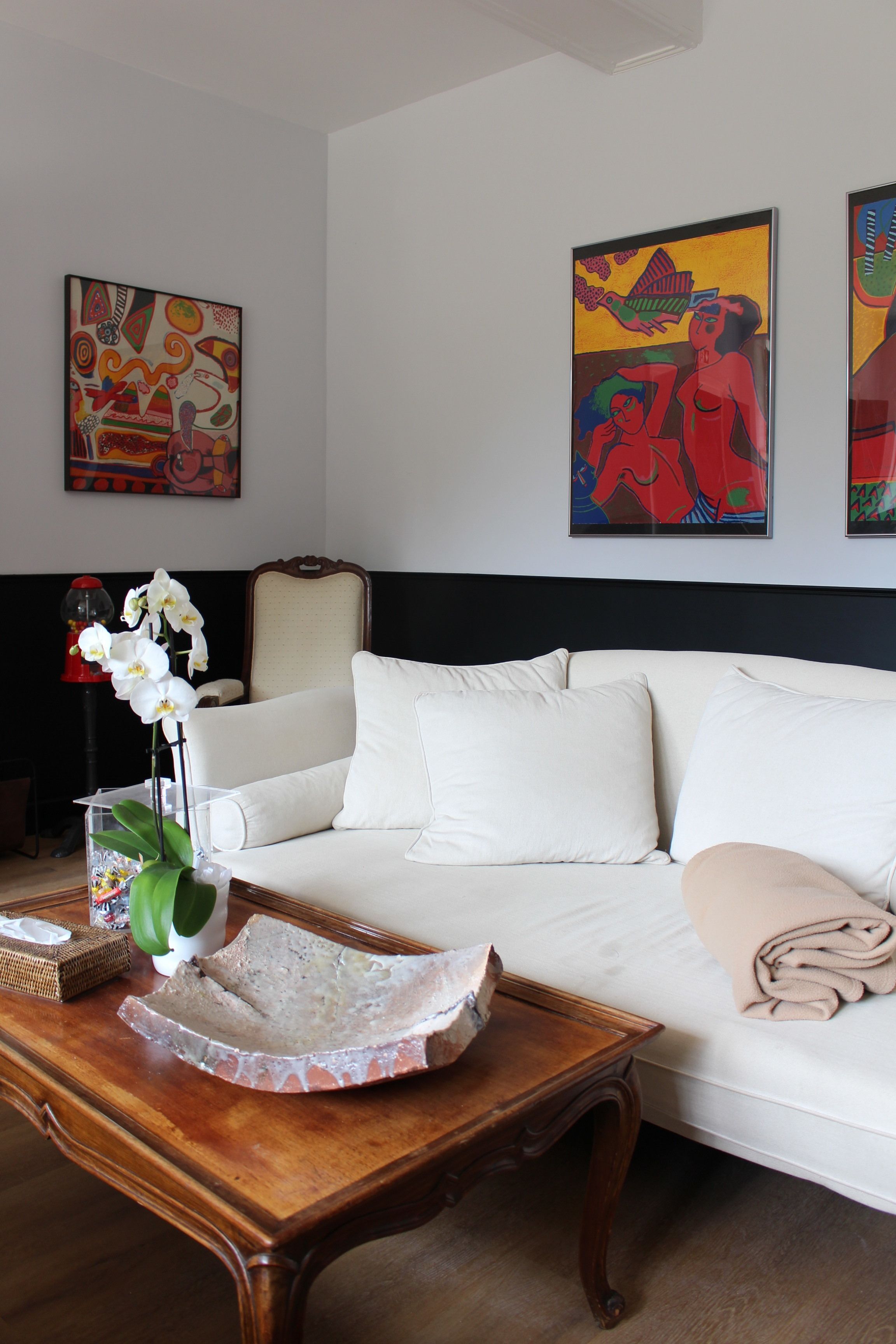 Un salon avec un canapé blanc, une table basse en bois et un vase rempli d’orchidées blanches. Des tableaux abstraits aux couleurs vives ornent le mur.