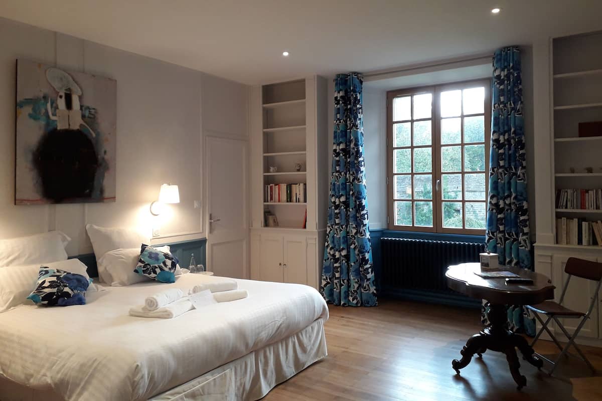 Una habitación de hotel con cama, escritorio, silla, estantería y ventana con cortinas.