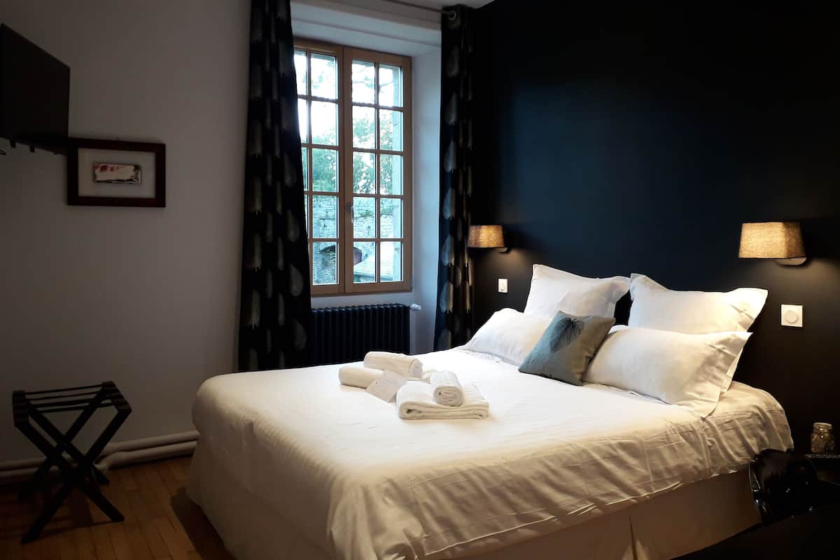 Una cama bien hecha con sábanas blancas y una toalla enrollada, una ventana con cortinas, una lámpara de pared y un cuadro en la pared.