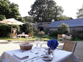 Un espace de restauration en plein air avec des tables disposées pour deux personnes, entouré d’une végétation luxuriante et avec un bâtiment en pierre en arrière-plan.