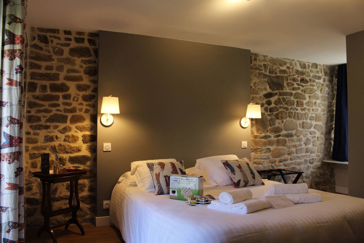 Una habitación de hotel con pared de piedra, cama con sábanas blancas, dos lámparas de noche y una mesita con una botella y un menú.