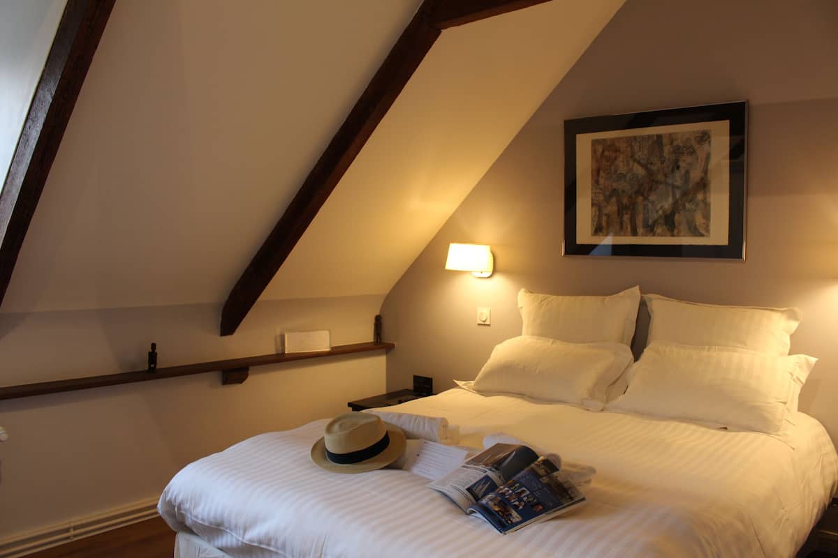 Una cama bien tendida con sábanas blancas, un sombrero y una revista sobre un piso de madera.
