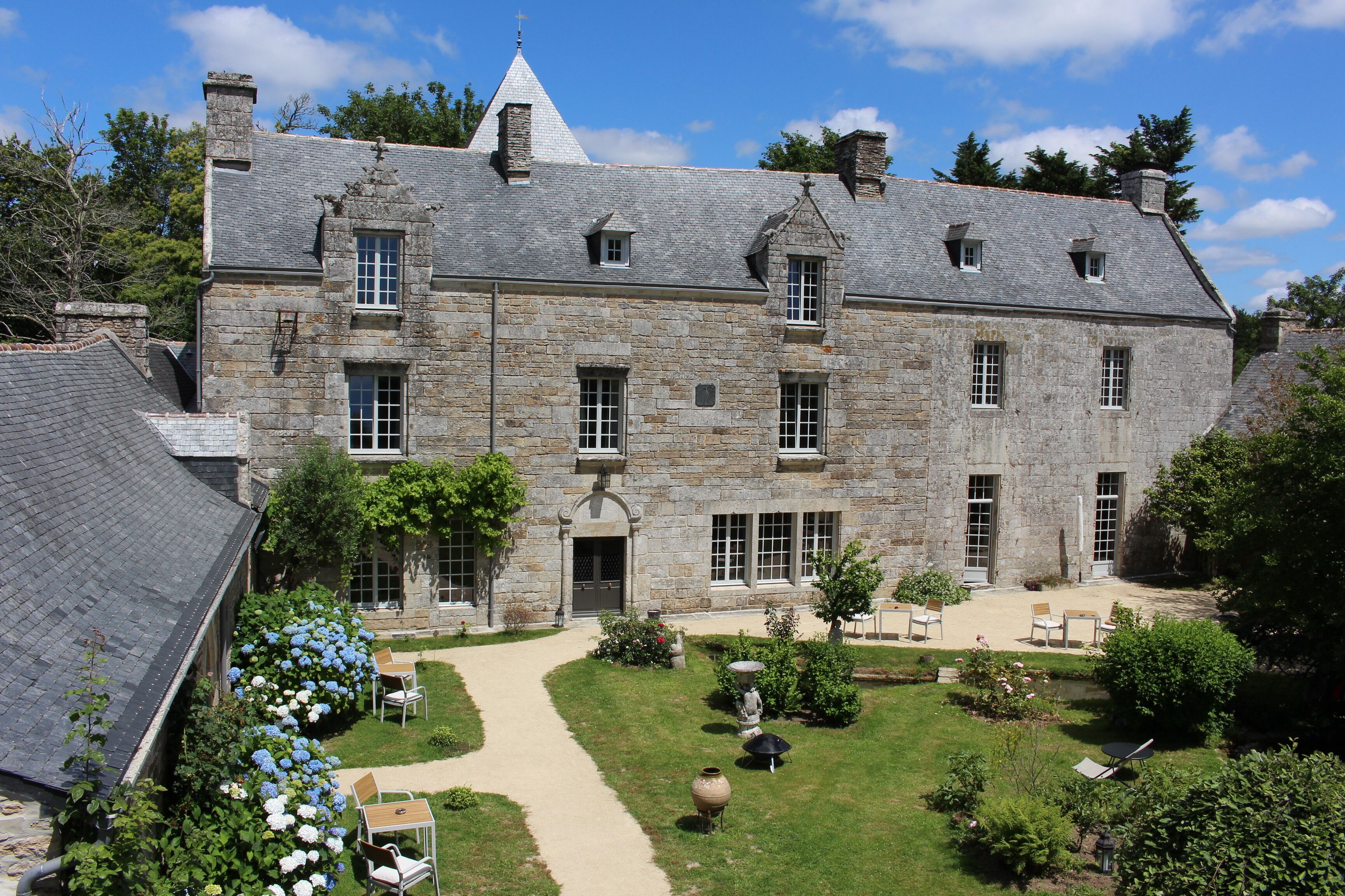 Un bâtiment en pierre doté d’une entrée centrale, de nombreuses fenêtres et d’un jardin aménagé avec des espaces pour s’asseoir à l’extérieur.