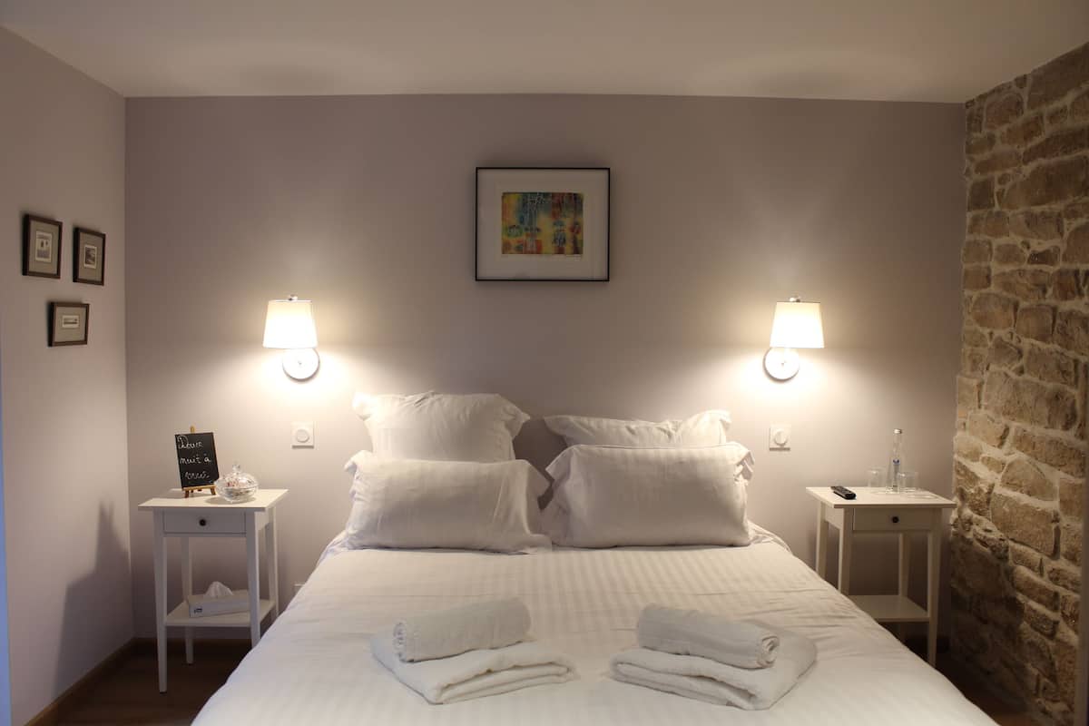 Una cama bien hecha con sábanas blancas, dos mesitas de noche con lámparas, un cuadro en la pared y una pared de piedra en un lateral.