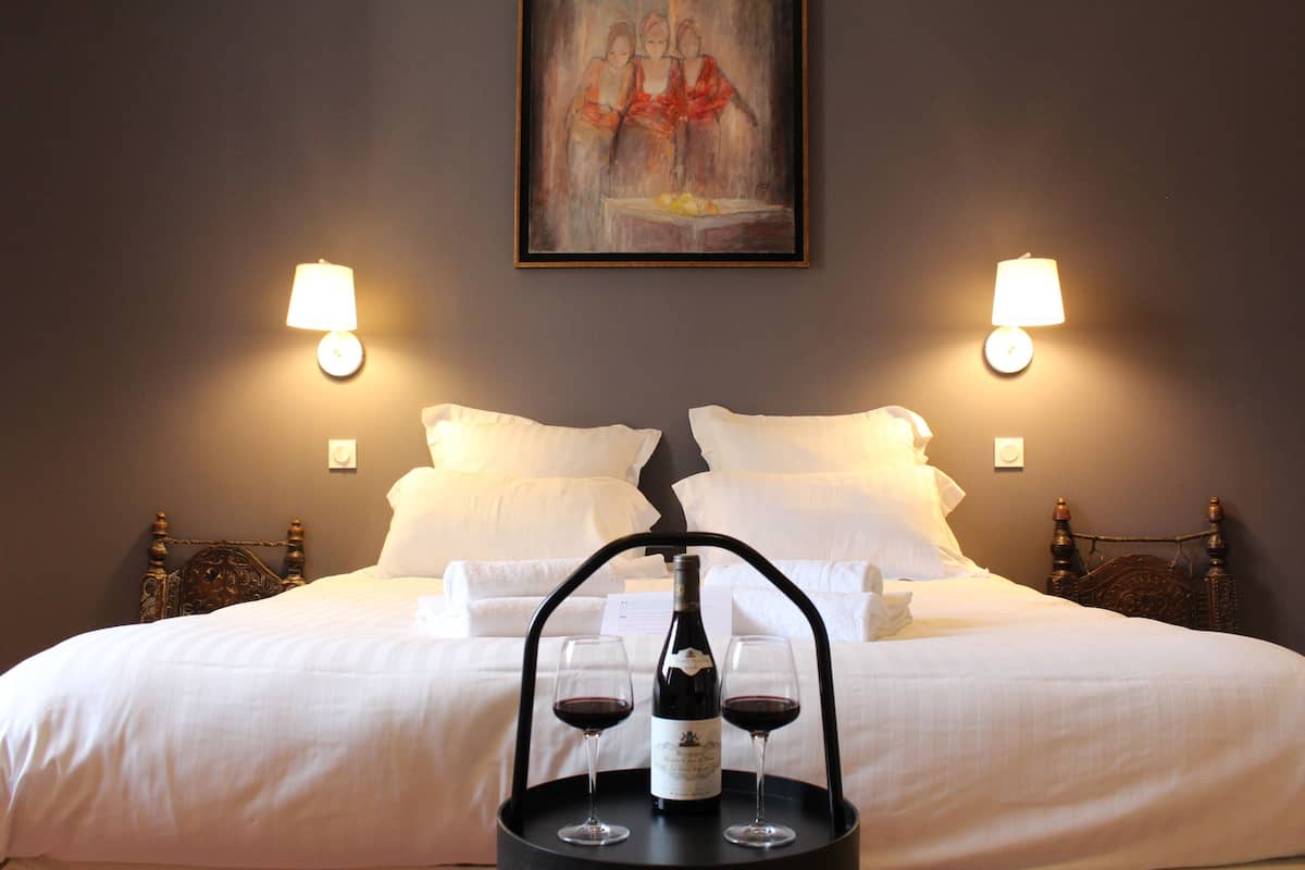 Una cama bien hecha con sábanas blancas, una botella de vino y dos copas de vino sobre una bandeja frente a ella.
