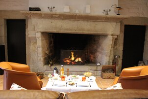 Un coin repas agrémenté d’une cheminée en pierre, de fauteuils en cuir, d’une table dressée pour deux personnes et d’un panier de pain.