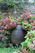 Un grand bac à fleurs en pierre, entouré d’une végétation luxuriante et de plantes d’hortensia.