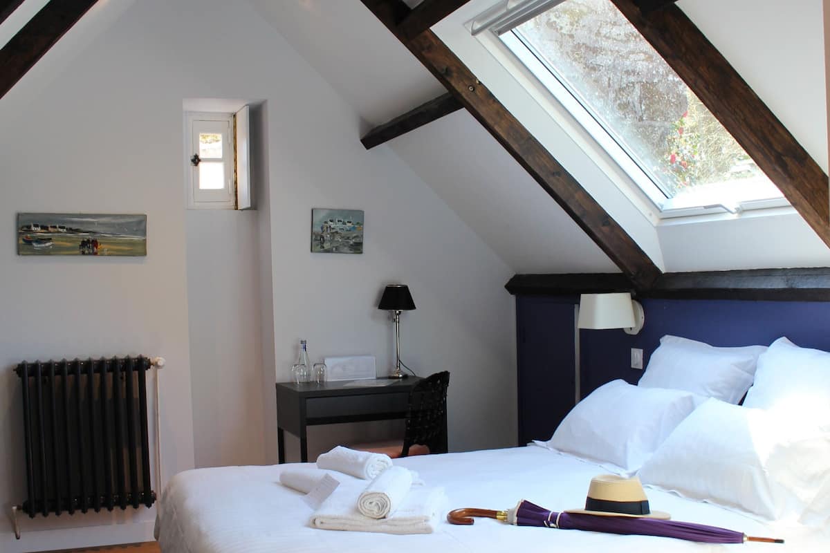 Un dormitorio con techo alto, vigas de madera, tragaluces, cama con sábanas blancas, mesita de noche con lámpara y radiador.