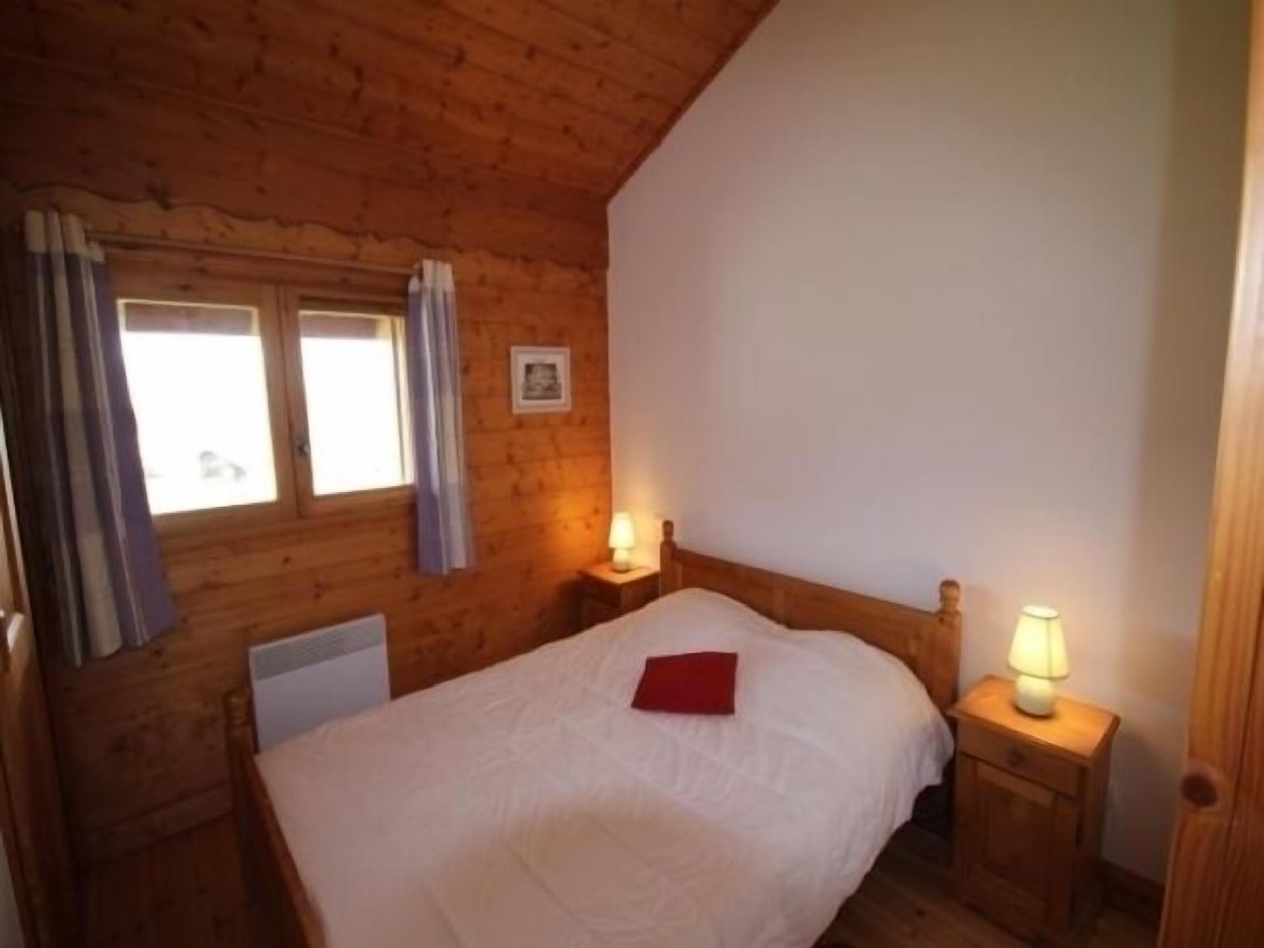 A wooden-paneled room with a bed, two bedside tables with lamps, a window with curtains, and a radiator.