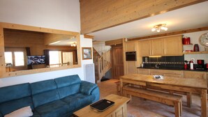 A cozy living room with a blue sofa, wooden coffee table, and a TV. Adjacent is a kitchen with wooden cabinets, a stove, and a dining table.
