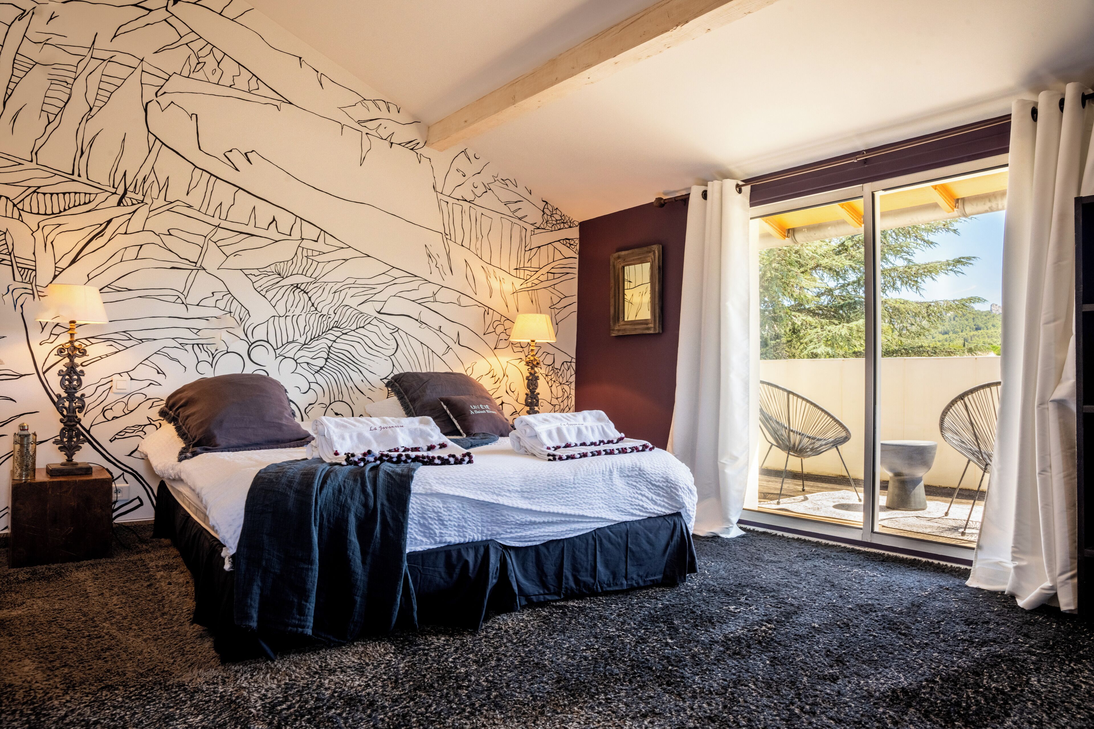 A bedroom with a large bed, a wall mural, a nightstand, a lamp, and a view of the outdoors through a sliding door.