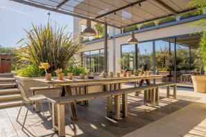 An outdoor patio with wooden benches and tables, surrounded by potted plants and a glass-enclosed building. - Villa Drossa, Magnificent family home near town center and Alpilles (Saint-Rémy-de-Provence)