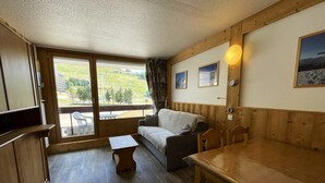 A cozy room with a wooden floor, a sofa, a small table, and a dining table. There is a window with a view of greenery and a building outside. - Renovated ski-in studio, 4 people, Les Menuires (Les Menuires)