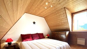 A cozy bedroom with a sloped ceiling, a bed with white bedding and red pillows, a nightstand with a red lamp, and a dresser. - Charming Breton House Steps from Saint-Guirec Beach, Ploumanac’h – 2 Beds, Garden, Pet-Friendly (Perros-Guirec)