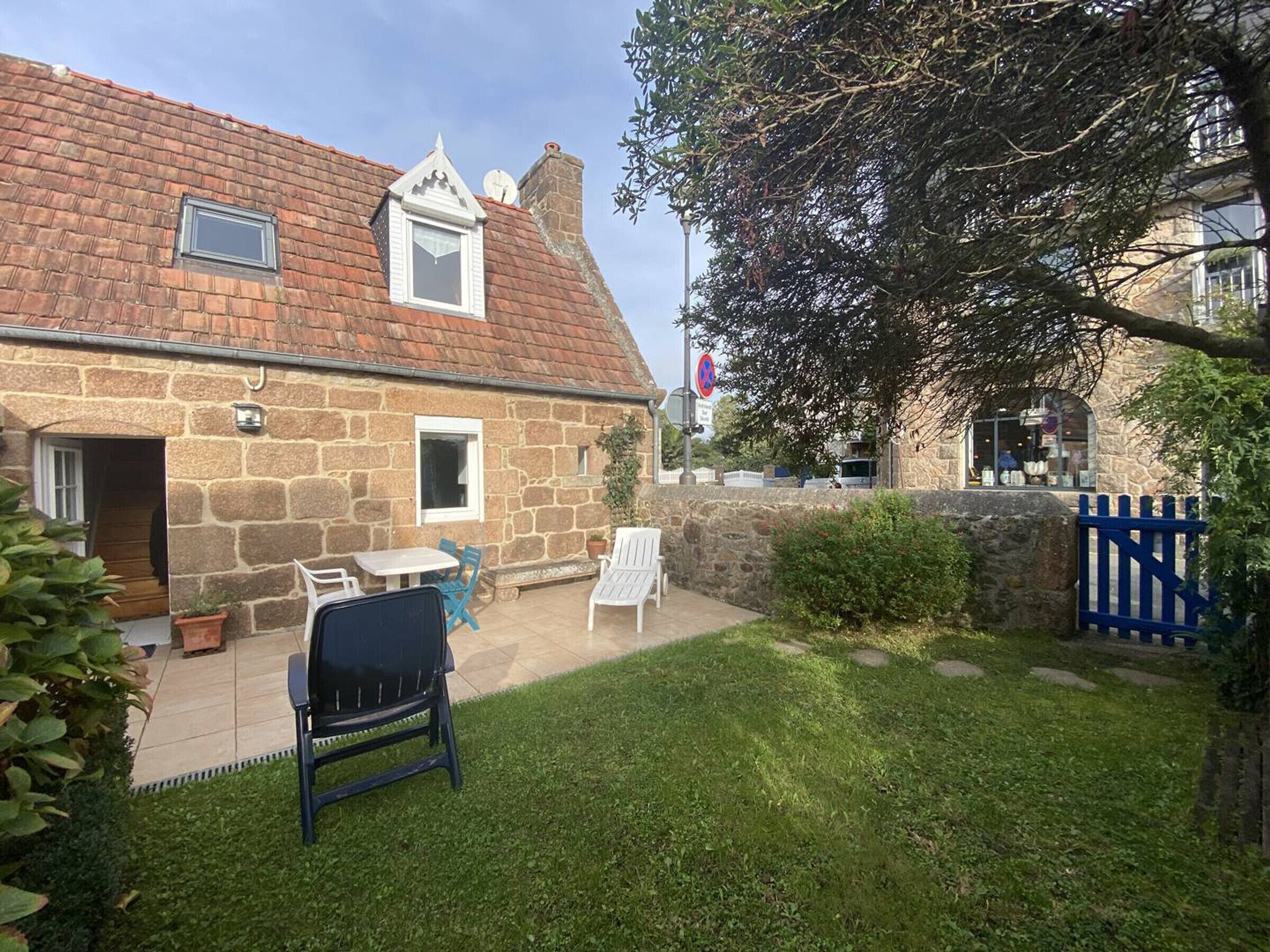 A stone house with a tiled roof, a small front porch with a table and chairs, and a blue gate.