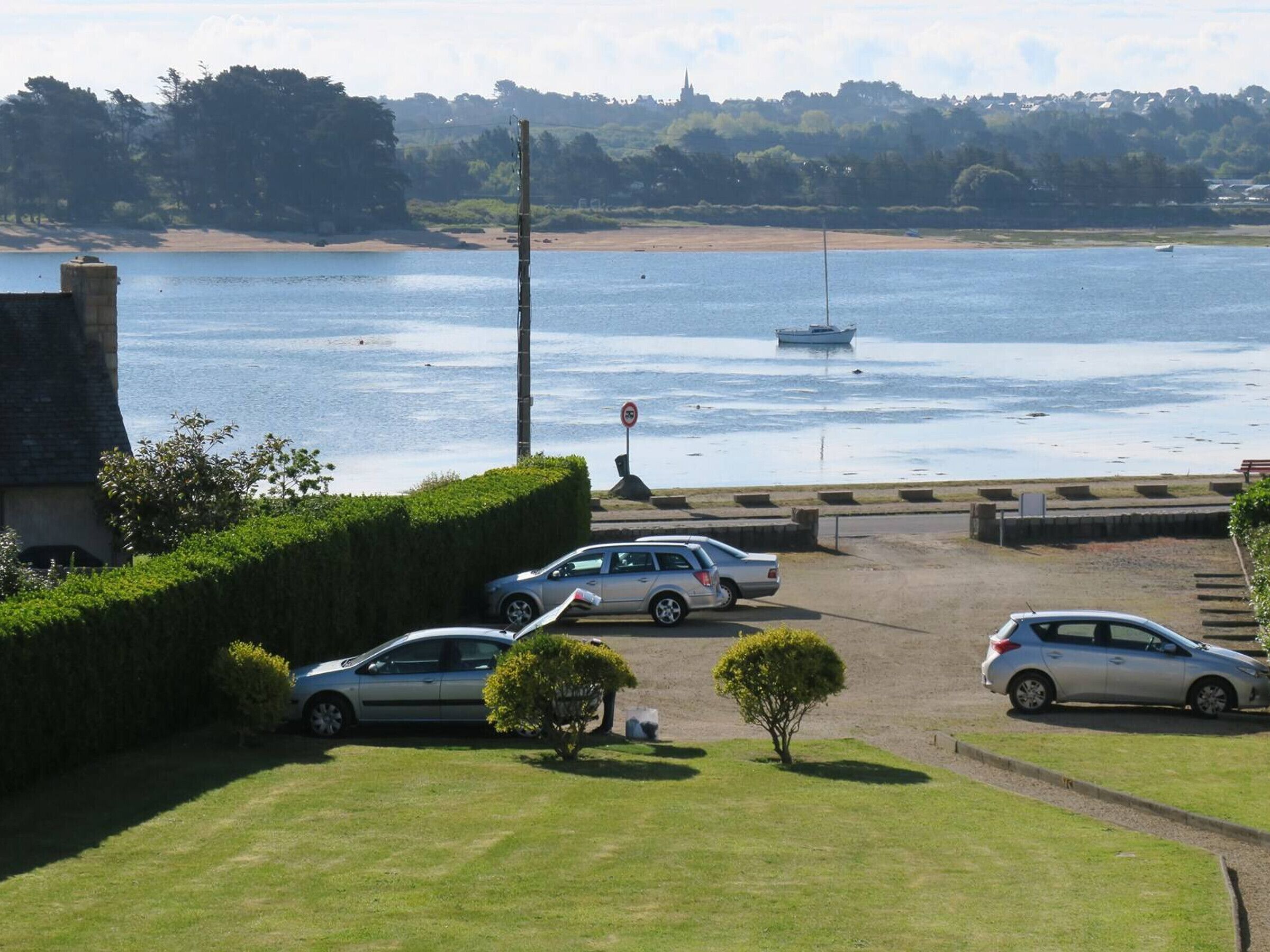A parking area with cars, a grassy foreground, and a view of a body of water with a boat in the distance.