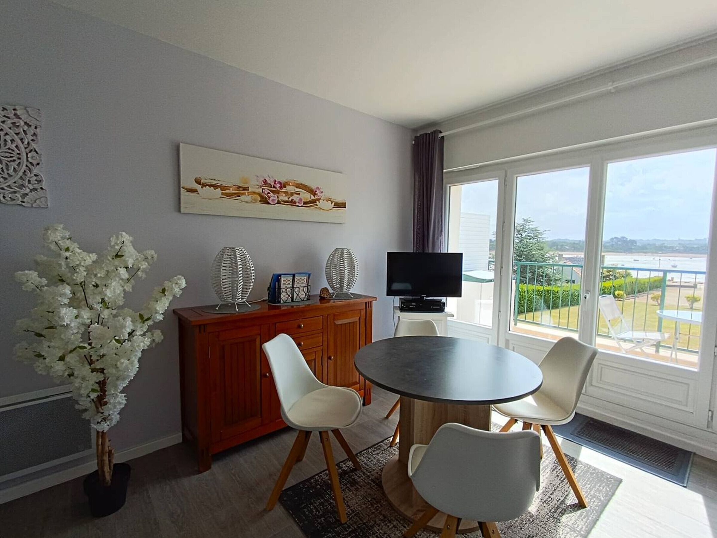 A modern living room with a round table, two white chairs, a wooden cabinet, a flat-screen TV, and a large window offering a view of a body of water and greenery.