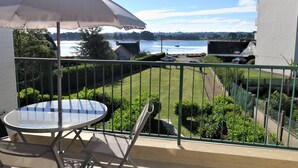 A balcony with a table and chairs, a table umbrella, and a view of a body of water and distant buildings. - Renovated apartment with terrace 200m from beaches in Trégastel (Trégastel)