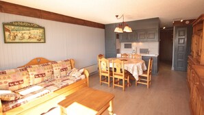 A cozy living room with a wooden sofa and a dining table, a kitchen area with blue cabinets, and a hallway leading to a door. - Les Saisies - 2-room with fireplace for 6 people (Les Saisies)