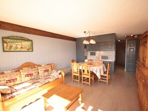 Ett mysigt vardagsrum med en soffa i trä och ett matbord, ett köksområde med blå skåp och en hall som leder till en dörr.