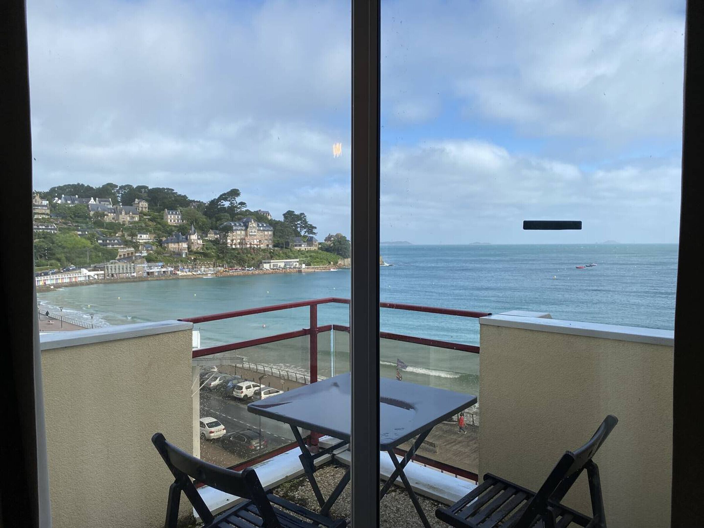 A balcony with a table and chairs overlooking a coastal town with buildings and a beach.