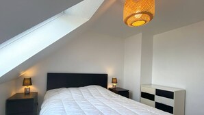 Un dormitorio con techo inclinado, una cama con edredón blanco, dos mesitas de noche con lámparas y un aparador blanco con cajones negros.