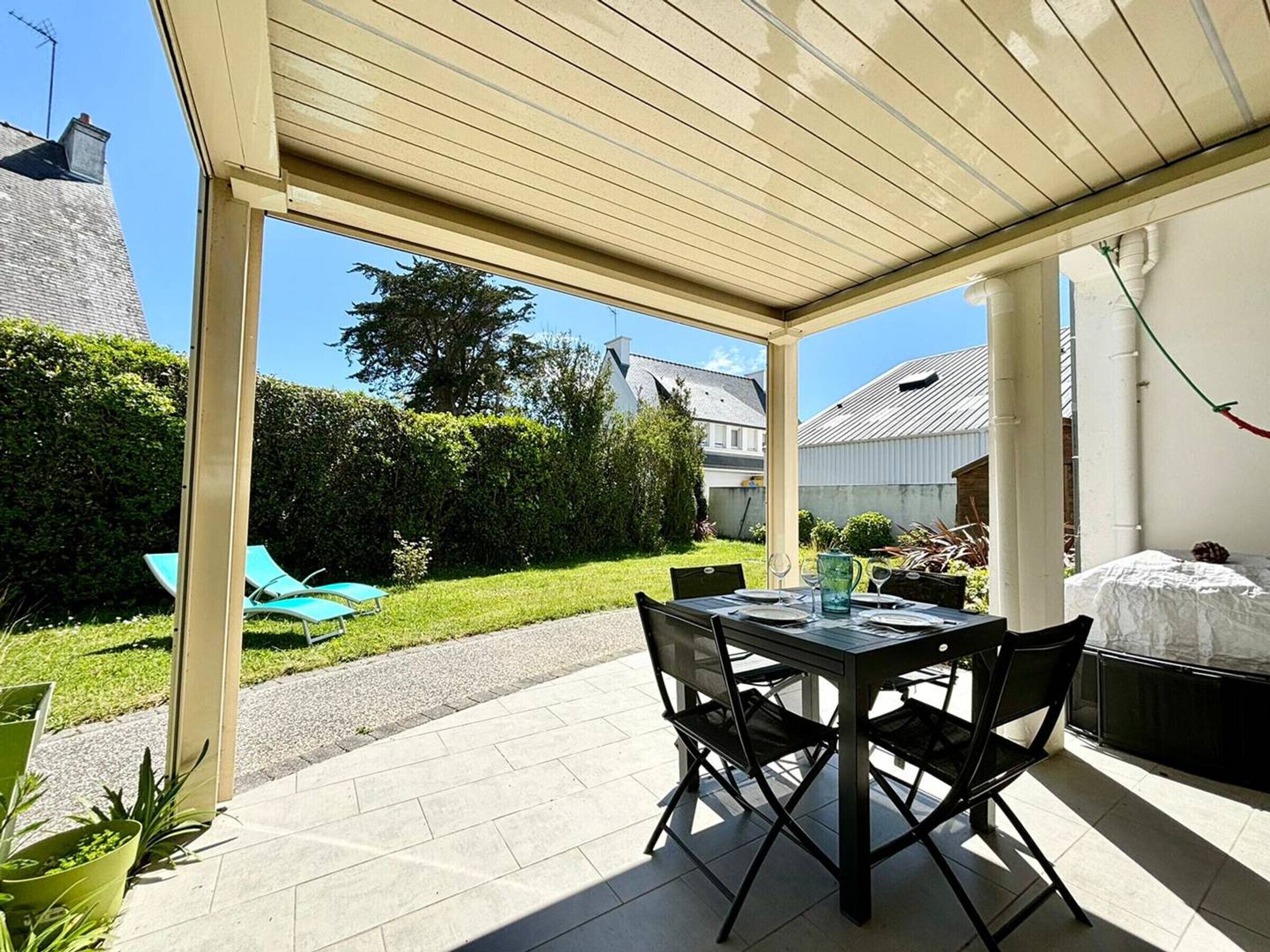 An outdoor patio with a table and chairs, a covered area, and a view of a garden and neighboring houses.