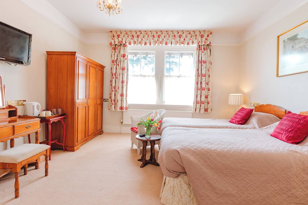 A bedroom with two beds, a wooden wardrobe, a desk with a chair, a small table with a vase of flowers, and a window with floral curtains.