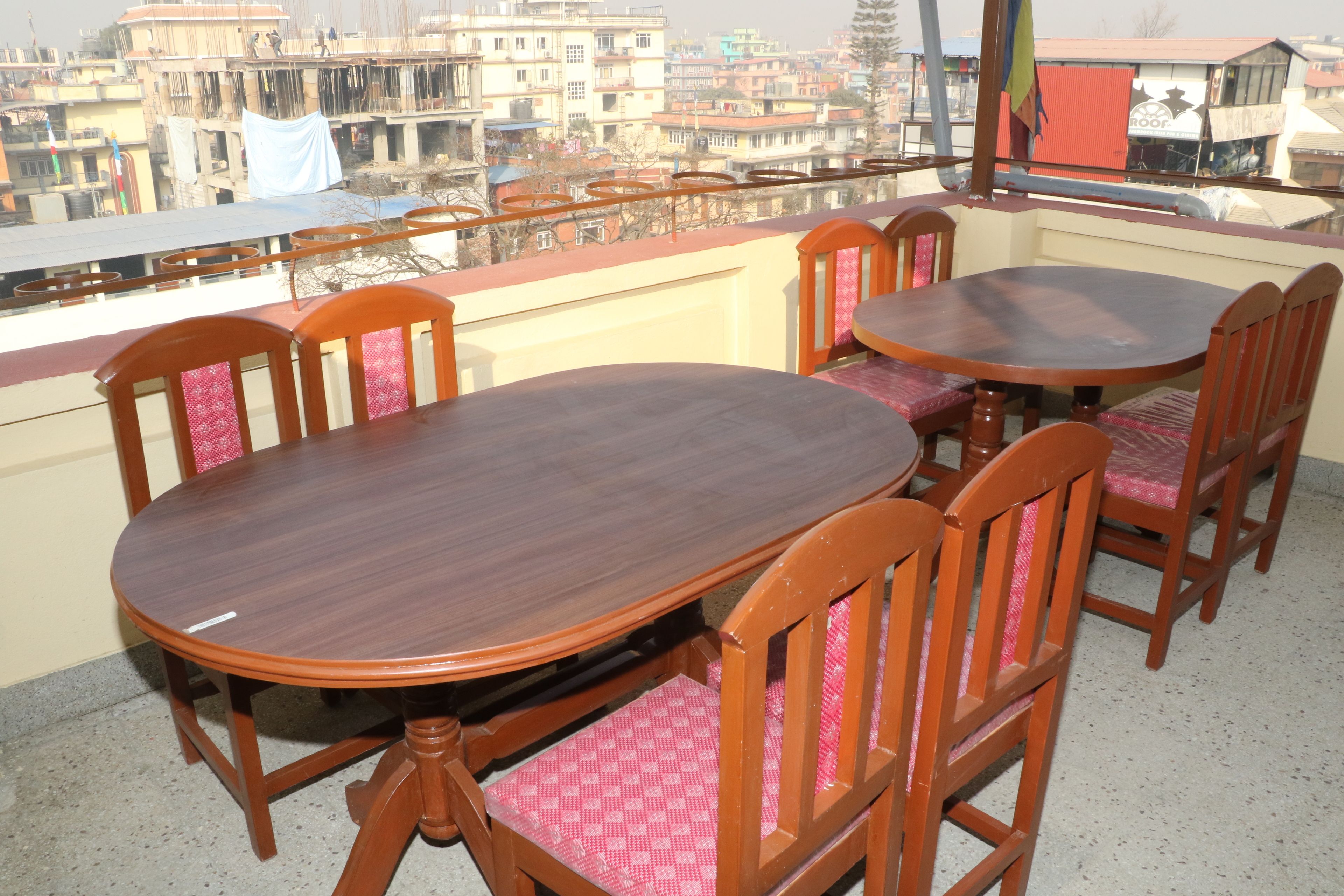 Un balcon avec des tables et des chaises en bois, donnant sur un paysage urbain composé d’immeubles et d’une structure surmontée d’un toit rouge.
