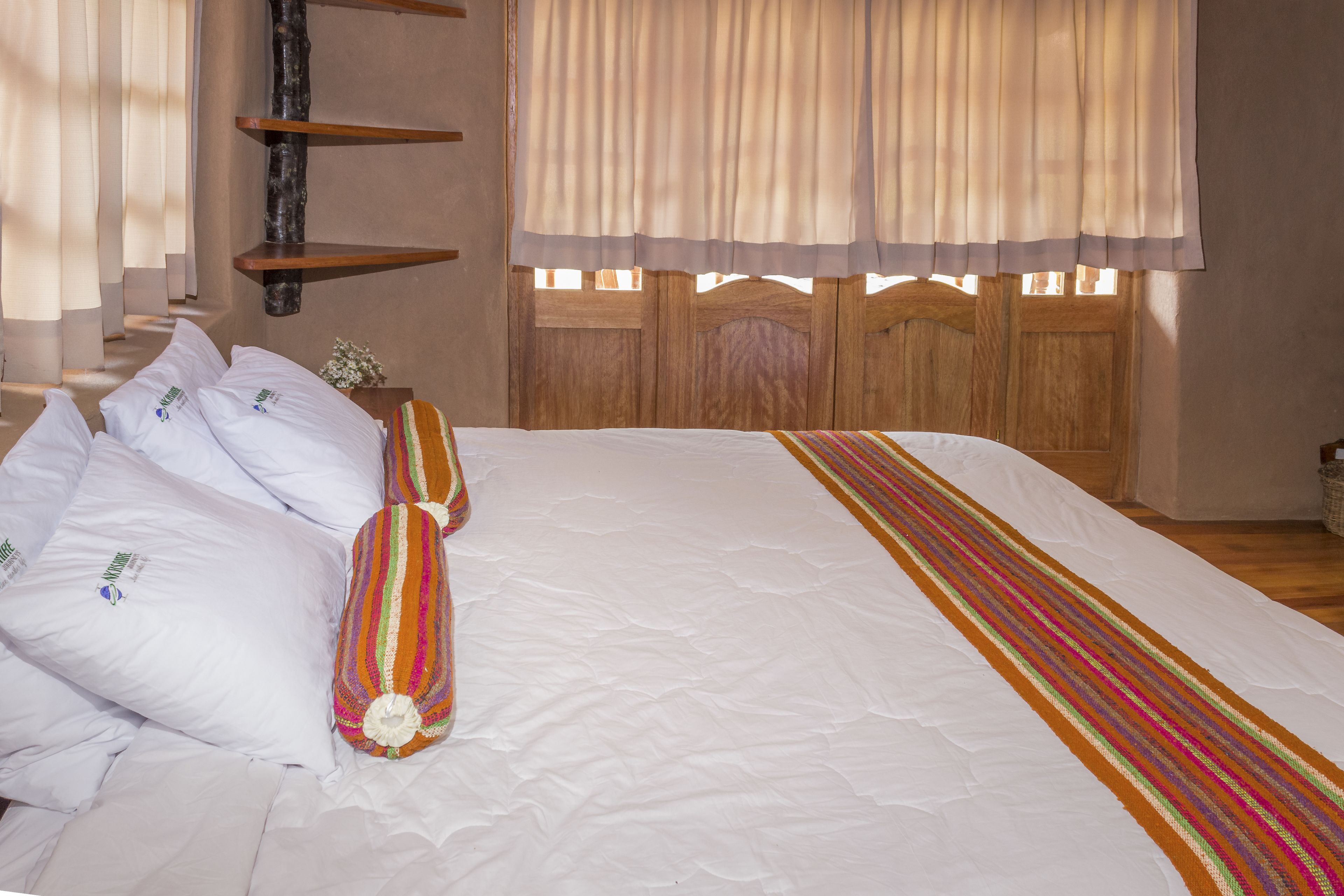 Una cama bien tendida con sábanas blancas y una manta a rayas de colores, cabecera de madera y una ventana con cortinas.
