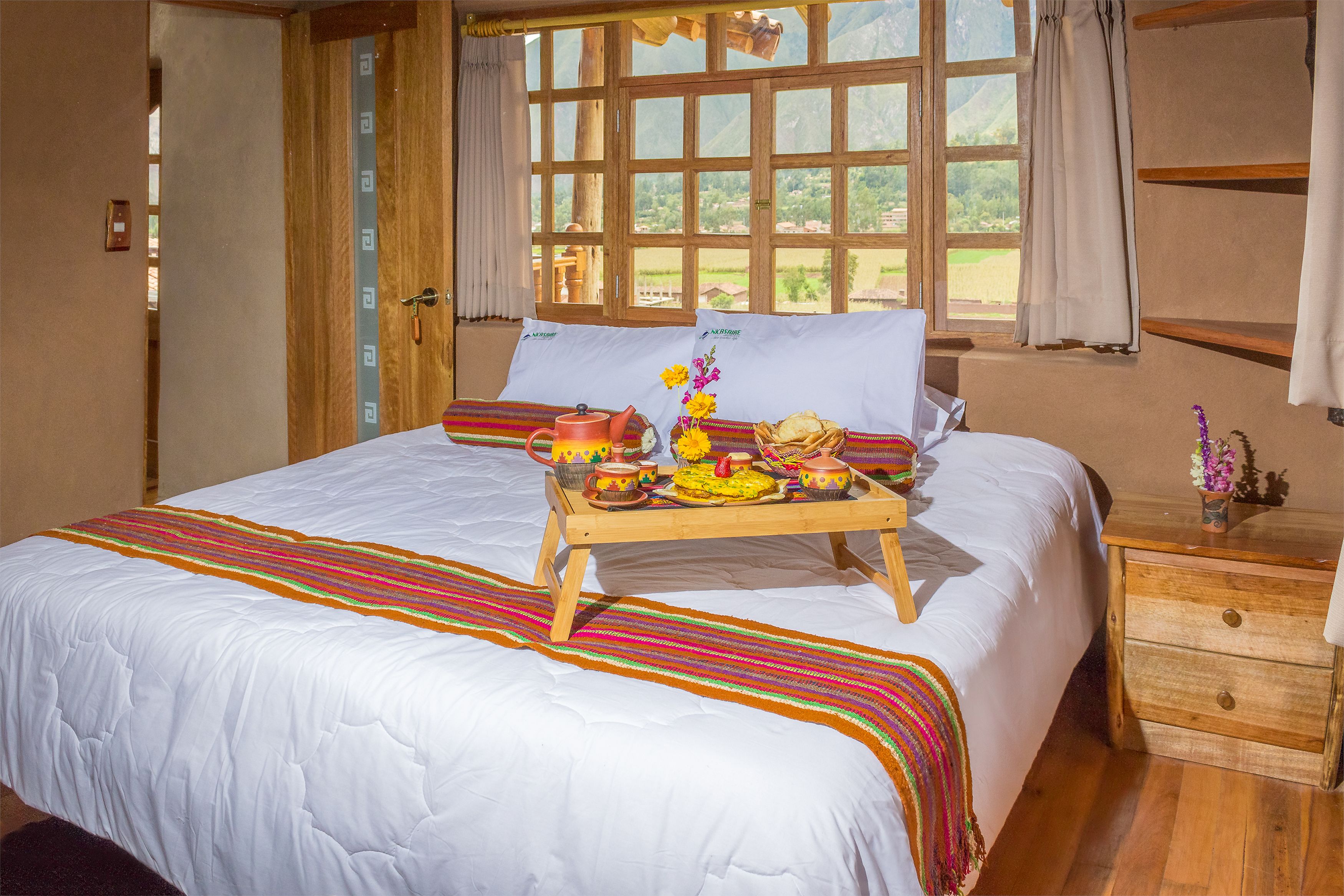 Una cama bien hecha con una bandeja de desayuno, una ventana con cortinas y una mesita de noche de madera con un cajón.