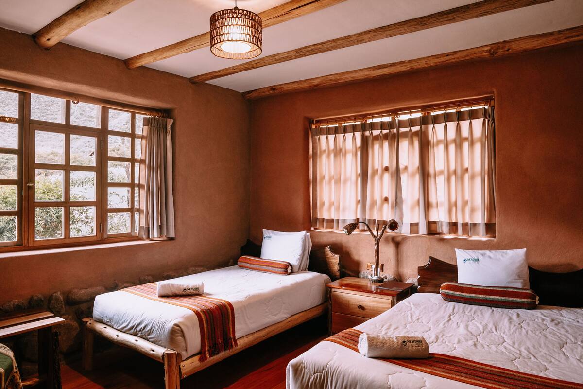 Una habitación con dos camas, vigas de madera, una pared de piedra y un ventanal con cortinas.