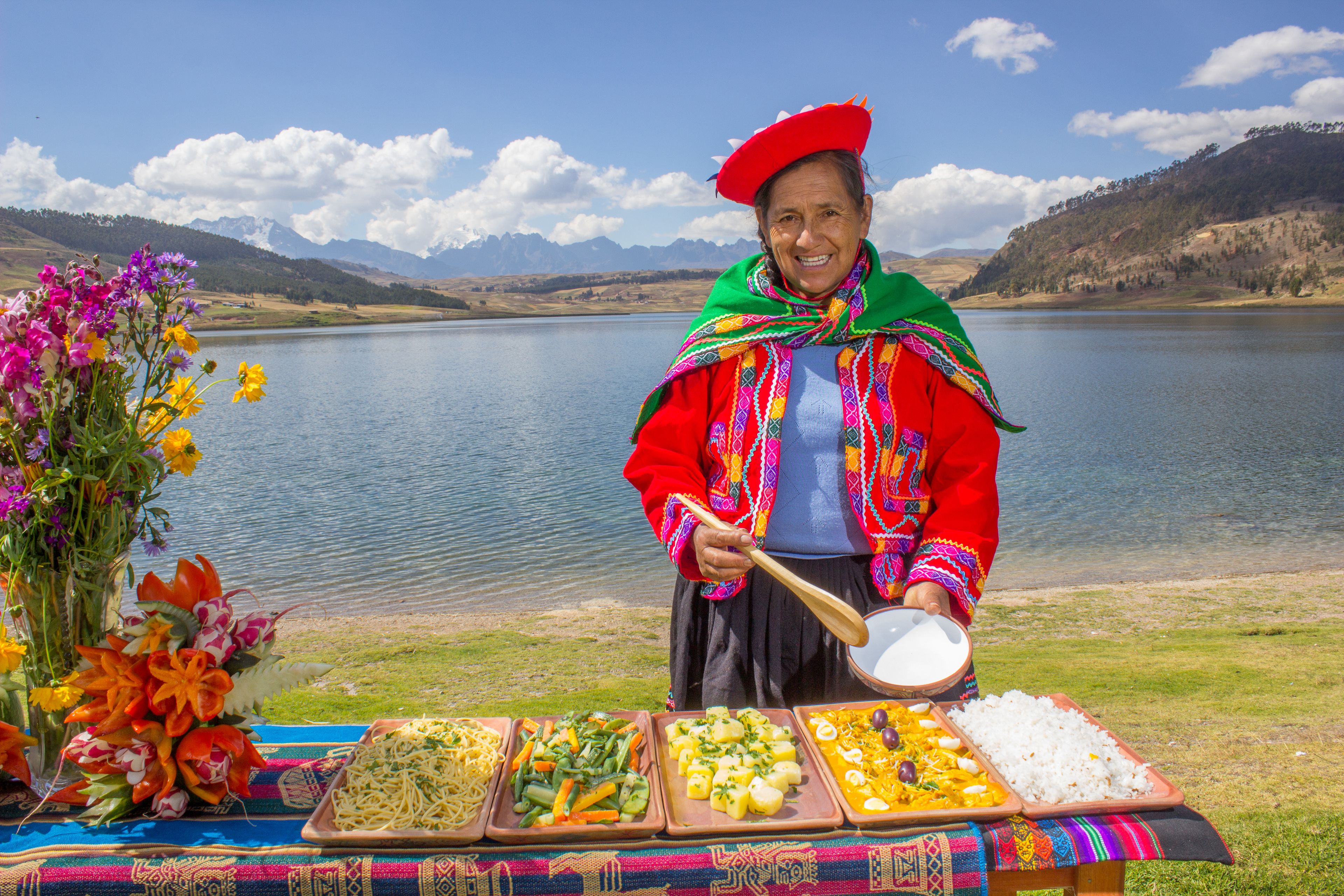A person serving food at a lakeside table with various dishes and a bouquet of flowers.