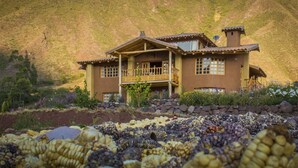 Una casa grande con balcón, rodeada de un jardín con diversas plantas y un campo de maíz al frente.