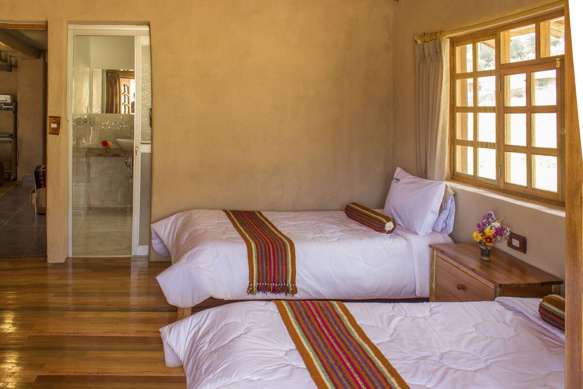 Habitación de hotel con dos camas, piso de madera, baño visible a través de una puerta abierta y una ventana con cortinas.